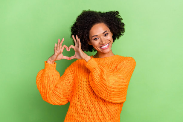 穿着橙色毛衣的甜美千禧一代卷发女士在绿色背景前做出心形手势的照片图片下载