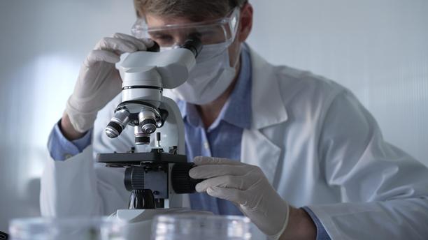 穿着白色手套、护目镜和医用口罩的男性科学家研究员正在现代实验室中使用显微镜，肖像视图。低调照明照片。科学与医学图片下载