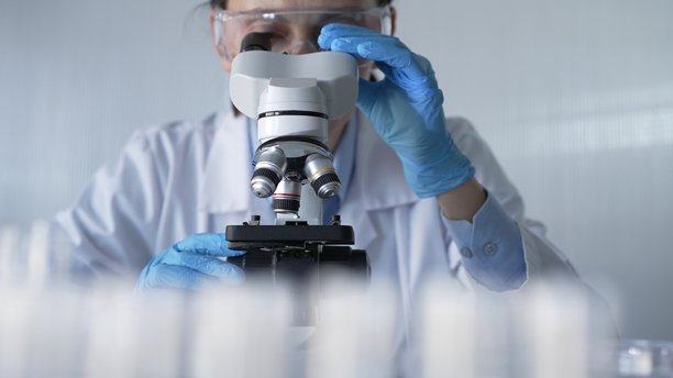 穿着实验室外套、戴着白色防护手套和眼镜的女科学家正在现代实验室中使用显微镜，肖像视图。低调照明照片。科学与医学图片下载
