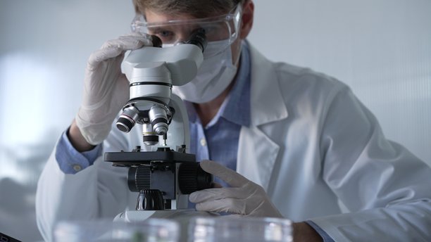 穿着白色手套、护目镜和医用口罩的男性科学家研究员正在现代实验室中使用显微镜，肖像视图。低调照明照片。科学与医学图片下载