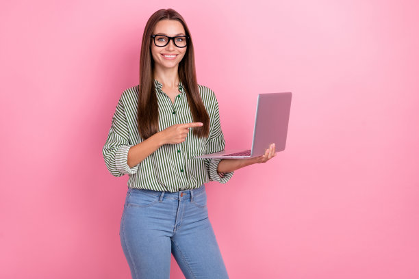 年轻的棕发女首席执行官戴着眼镜，穿着衬衫和牛仔裤，坐在粉色背景前使用笔记本电脑的照片图片下载