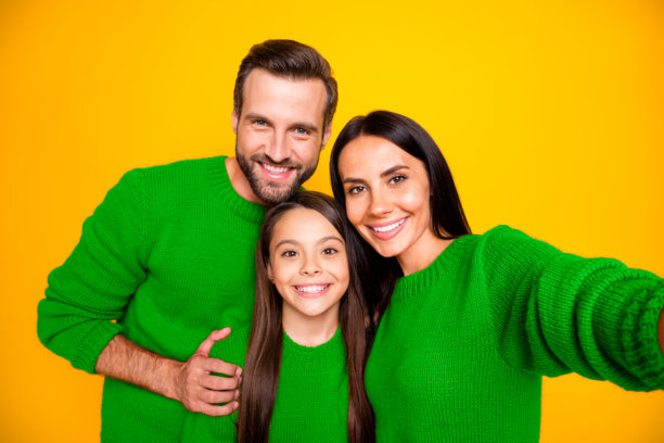 有趣、快乐、可爱的家庭照片，妈妈在自拍，爸爸抱着穿红色毛衣的女儿，大家一起在鲜艳的黄色背景下。图片下载