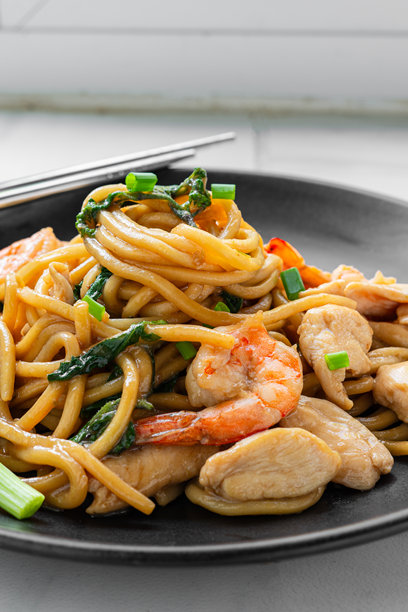 美味的炒乌冬面，搭配罗勒和干辣椒。用蚝油、鸡胸肉和虾炒制，最后用葱花装饰，摆放在黑色盘子上。家庭厨师的竖版照片图片下载