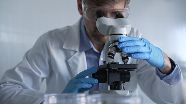 穿着蓝色手套和防护眼镜的男性科学家研究员正在现代实验室中使用显微镜，肖像视图。低调照明照片。科学与医学概念。图片下载