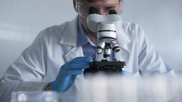 穿着蓝色手套和防护眼镜的男性科学家研究员正在现代实验室中使用显微镜，肖像视图。低调照明照片。科学与医学概念。图片下载