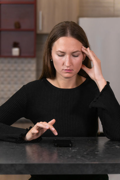 年轻女性坐在厨房桌旁，手握智能手机，因工作压力感到沮丧和疲惫，双手托着头。竖版照片图片下载