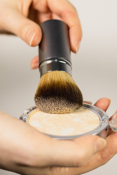 垂直照片，使用散粉。女人正在用可伸缩的面部刷轻拍粉饼。她用刷子沾取粉末，准备涂抹在脸上。图片下载