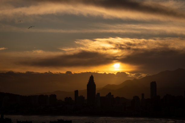 西班牙贝尼多姆老城的黄昏日落照片，展现了美丽的红色日落和天空中的云彩，以及夏季高层公寓的景象。图片下载