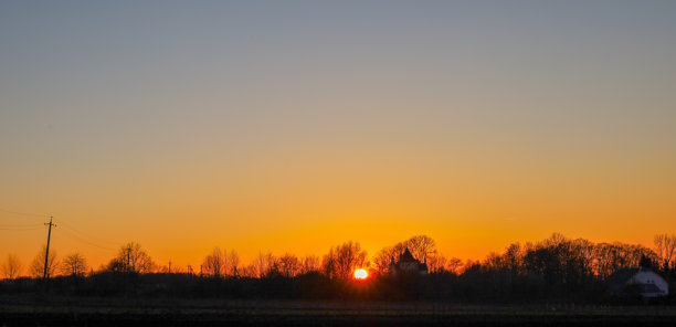 这张照片捕捉了乡村风景下迷人的日落。夕阳在树木的轮廓后落下，散发出明亮的橙色光芒，逐渐过渡到天空上方柔和的蓝色调。图片下载