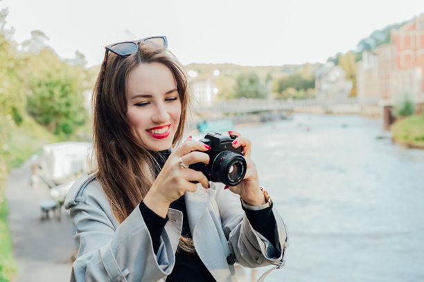 微笑的年轻女性手持复古风格相机。女性旅行者在城市漫步时查看相机中的照片。旅游博主、摄影师、网红捕捉瞬间。爱好，旅行概念。图片下载