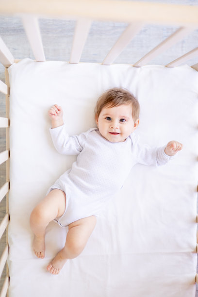一个小孩在家里的白色棉床上，穿着白色连体衣，微笑着躺在床上，六个月大的小女孩，眼睛大大的，俯视图。图片下载