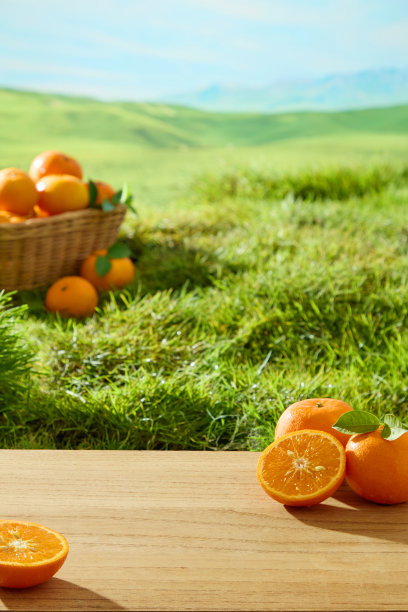 自然风光的竖版照片，藤编篮子里装着成熟的橙子，放在绿色草地上。新鲜的橙子被切成两半，露出多汁的果肉。远处可见山脉。图片下载