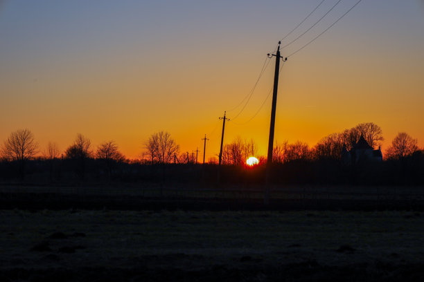 这张照片展示了一个田野上的日落，电线穿过画面。太阳在树木的后面落下，照亮了天空，呈现出明亮的橙色和黄色。图片下载