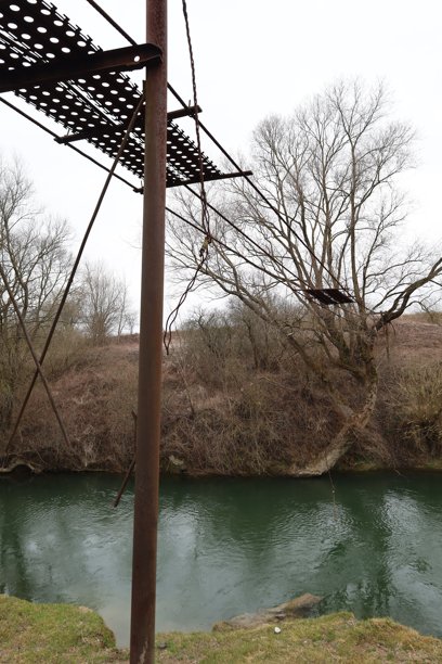 这张照片展示了一条水呈绿色的河流，前景中可见一个带有穿孔元素的金属结构。该结构呈现出生锈的棕色调。图片下载