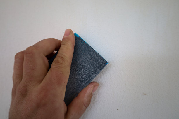 这张照片是一个人手握砂纸在打磨白色墙壁的特写。砂纸是深灰色的，表面粗糙。图片下载