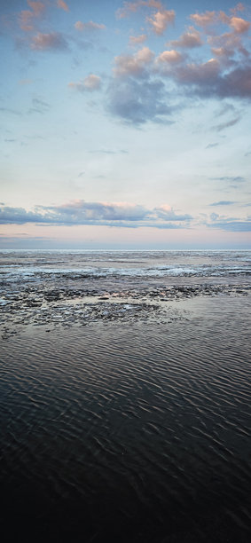 春季风景。美丽的海湾晨曦全景。明亮的光线下，水面上漂浮着积云。海岸线上有冰雪和岩石。阳光的光芒在海面上反射。竖版照片图片下载