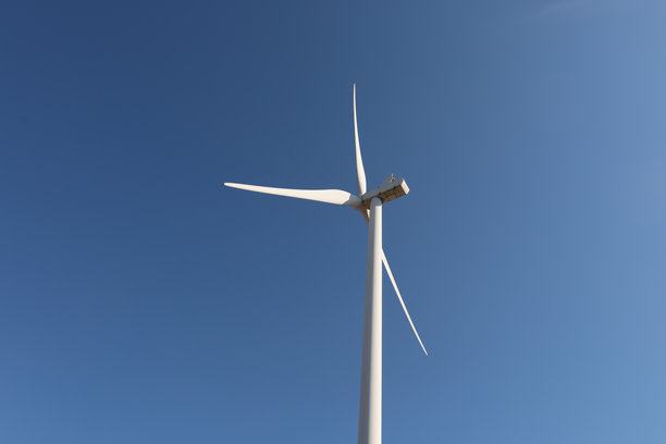 一座风力涡轮机矗立在明亮的蓝天之下。风力涡轮机的塔身和叶片都是白色的。这张照片传达了清洁能源和现代科技的感觉。图片下载