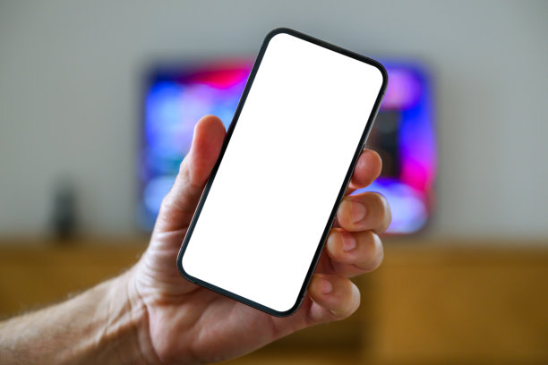 智能手机模型屏幕，男子在电视机前手持手机，电视遥控应用和电视节目列表概念图片下载