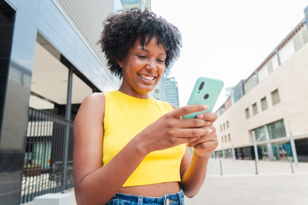 非裔美国女孩微笑着使用手机浏览互联网应用。青少年女孩用手机在社交媒体上分享照片。年轻女性在户外拿着智能手机。图片下载