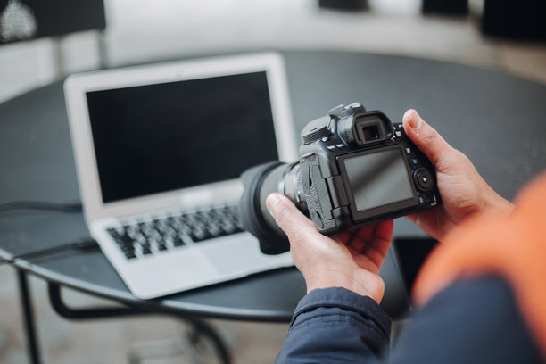 摄影师在拍摄后查看相机显示屏上的照片，桌子上放着准备编辑的笔记本电脑。图片下载