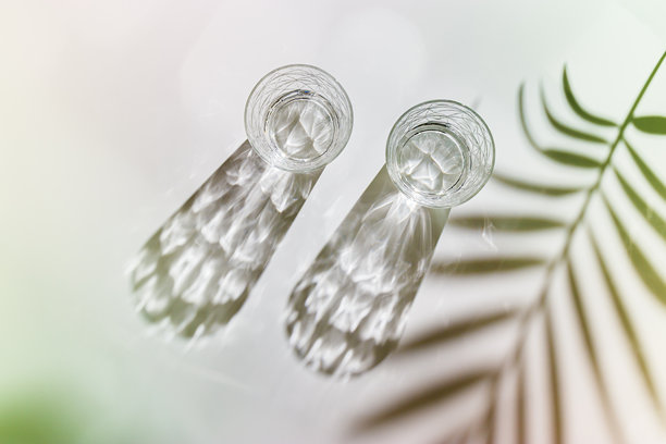 白色背景上的水杯，棕榈叶的阴影和阳光的反射，彩色照片，极简风格，夏季静物的美学俯视图，透过水晶杯的折射，光晕效果。图片下载