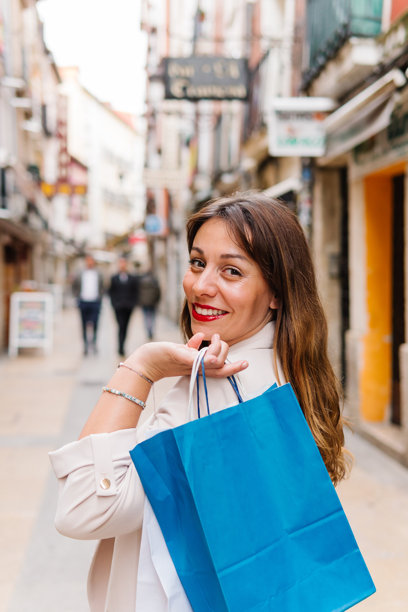 快乐的年轻女性涂着红色口红，手提购物袋，转身微笑着看向镜头，站在商业街上。图片下载