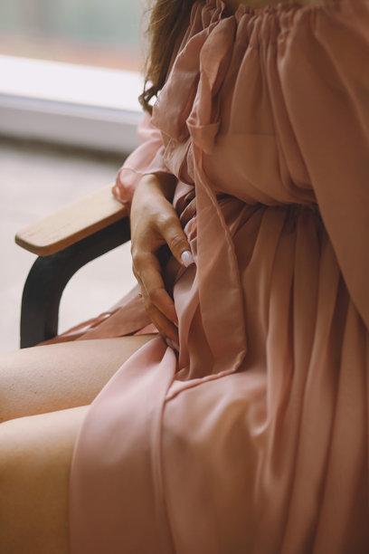 怀孕早期的女性特写照片，穿着时尚的粉色睡袍，坐在椅子上，手托着肚子，室内拍摄。图片下载