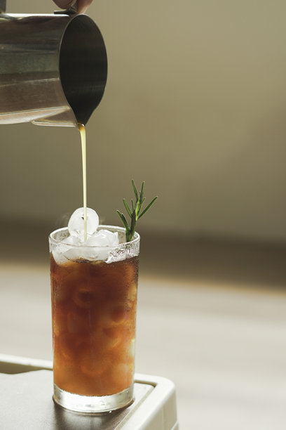 手握壶倒出浓缩咖啡，倒入透明玻璃杯中，杯内有冰块和橙汁，点缀着迷迭香叶。这是一款清爽的夏季饮品菜单项，竖版照片。图片下载