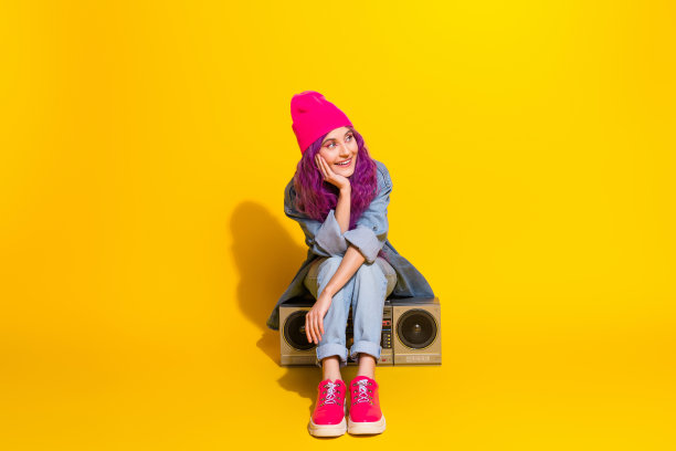 年轻女性坐在音响上，穿着鲜艳的粉色鞋子和毛线帽，背景是明亮的黄色，展现出色彩斑斓的时尚照片。图片下载