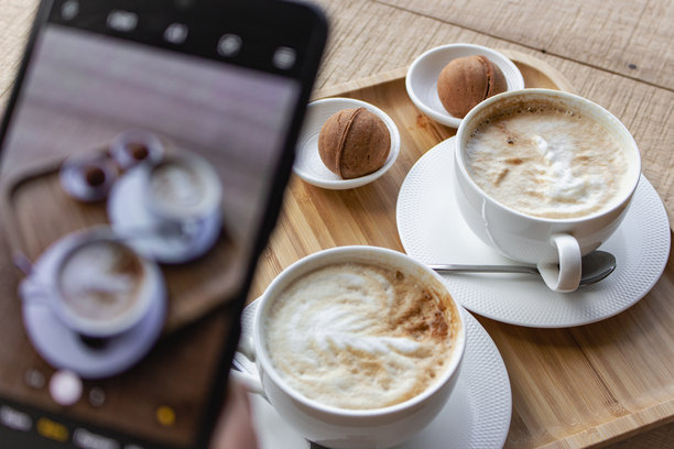 在手机屏幕上拍摄两只白色咖啡杯和甜点的照片。早餐平铺在手机屏幕上。拍摄早晨香气四溢的咖啡和甜点，适合社交媒体。屏幕上有美式咖啡加牛奶和饼干。图片下载