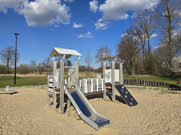 这张图片描绘了一个游乐场，里面有一个木制游乐设施，包括滑梯，位于沙地上。背景中有树木和蓝天白云。图片下载