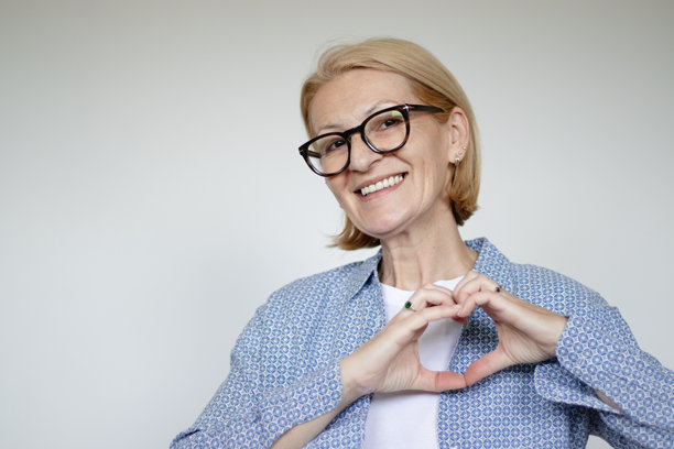 成熟的金发女性戴着眼镜，在白色背景前用双手做出心形手势的工作室拍摄照片图片下载