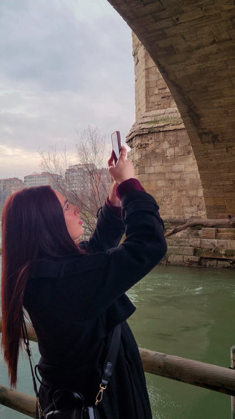年轻女性在西班牙萨拉戈萨的一座历史石桥下拍摄城市风景和河流的照片图片下载