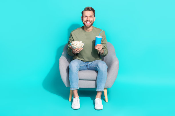 年轻的金发男孩穿着毛衣、牛仔裤和运动鞋，坐在青绿色背景下看电视的全身照片图片下载