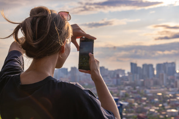 女人在日落时用智能手机拍摄城市天际线。她站在屋顶上，背景是城市。照片是从背面拍摄的。图片下载