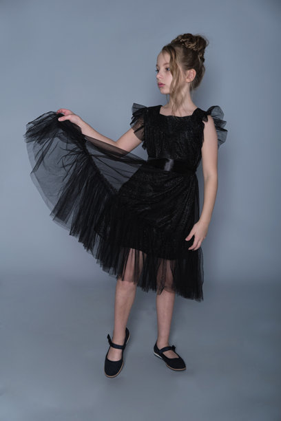 一个女孩优雅地站在一件黑色荷叶边裙子里，轻轻提起裙子，展现出精致的薄纱层。照片强调了优雅和气质，突显了儿童正式服装的现代趋势。图片下载