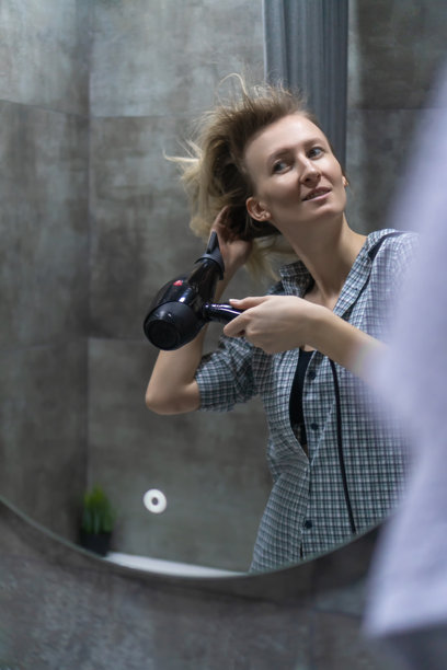 女性在光滑的浴室镜子前用吹风机吹干头发，体现了现代家庭环境中日常生活和个人护理的本质。竖版照片图片下载