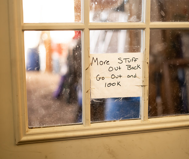 一张特写照片，展示了一张手写的标志，上面写着“后面有更多东西，出去看看”，贴在一扇脏污的玻璃门内侧。背景模糊，似乎是一个车库或储藏区。图片下载