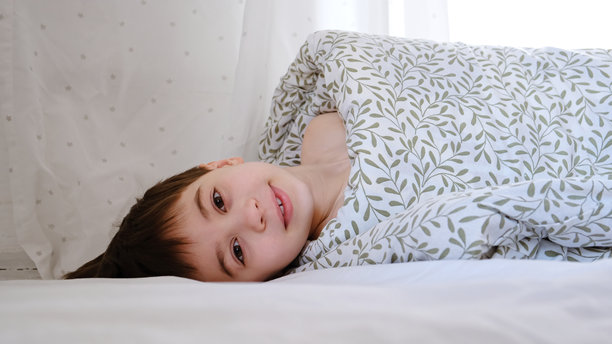 卷起的人。一个裹着毯子的孩子，头部露在外面，躺在卧室的床上。阳光明媚的早晨梦境。心理上的安慰。快乐微笑的白人男孩。高质量照片。图片下载