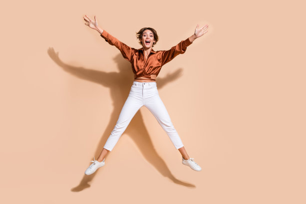 穿着棕色上衣的活泼积极女士在米色背景下高高跳起，像一颗星星一样，展现出全身的照片。图片下载