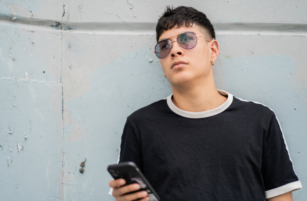戴着太阳镜和智能手机的沉思年轻人靠在城市墙壁上 - 股票照片。图片下载