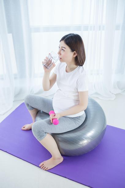 亚洲孕妇坐在健身球上做瑜伽，手持哑铃，喝水。图片下载