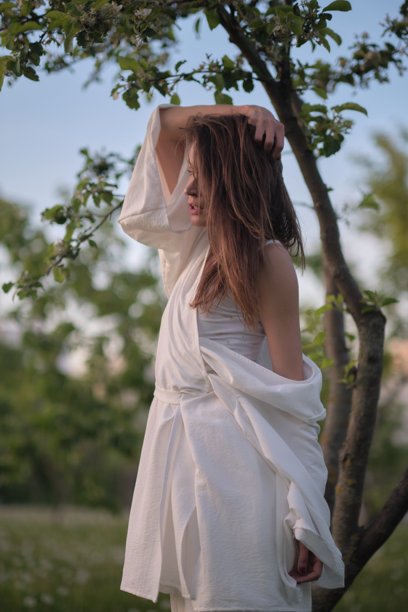 一位穿着白色裙子的女性站在树旁，微笑着回头看。这张图片捕捉了在轻松的户外环境中与自然的快乐和联系。图片下载