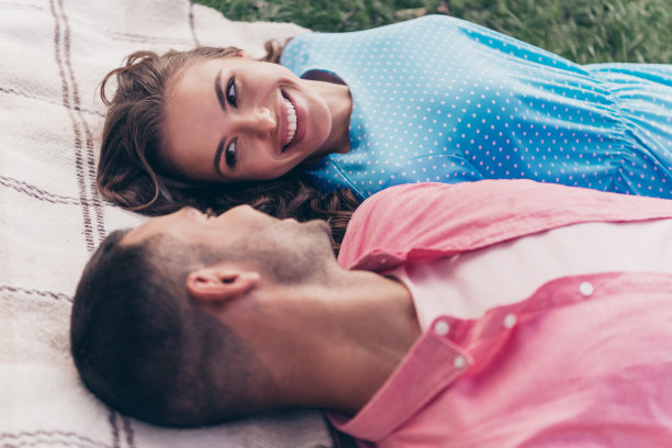高角度拍摄的照片，展示了一对穿着夏季服装的年轻夫妻在户外后院的格子毯子上放松，面带微笑。图片下载