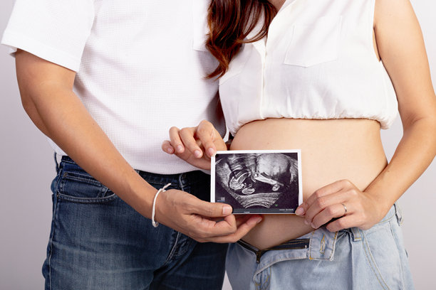 快乐的亚洲家庭夫妇在白色背景下，丈夫拥抱着妻子，妻子则在肚子上拿着超声波婴儿照片。美丽的孕妇母亲，肚子很大。图片下载