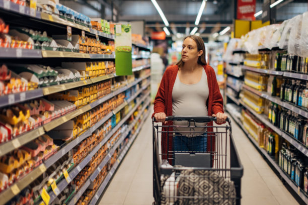 一位孕妇推着购物车，走过货架间，注视着超市货架上展示的食物。图片下载