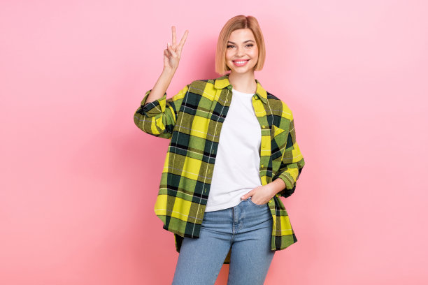漂亮年轻女孩的照片肖像，展示V字手势，打招呼，穿着格子黄色服装，背景为粉色。图片下载
