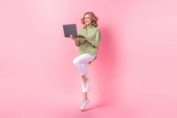 穿着绿色衬衫的闪亮甜美女性在粉色背景下跳得很高，正在用现代设备发送电子邮件的全身照片图片下载