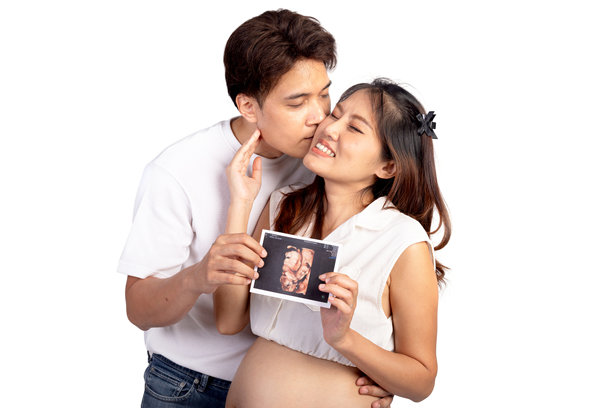 快乐的亚洲家庭夫妇在白色背景下，亚洲丈夫亲吻他美丽的妻子，同时他们手里拿着超声波婴儿照片。美丽微笑的孕妇母亲，肚子隆起。图片下载