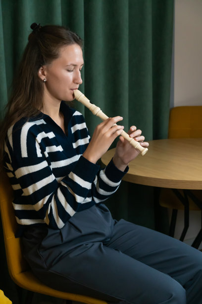 年轻女性音乐家舒适地坐在椅子上，在家中演奏直笛，专注于完善她的木管乐器技巧，同时享受创作的过程。竖版照片图片下载